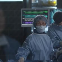 two people standing near the patient in the operating room, one stands straight and looks forward, other does surgery