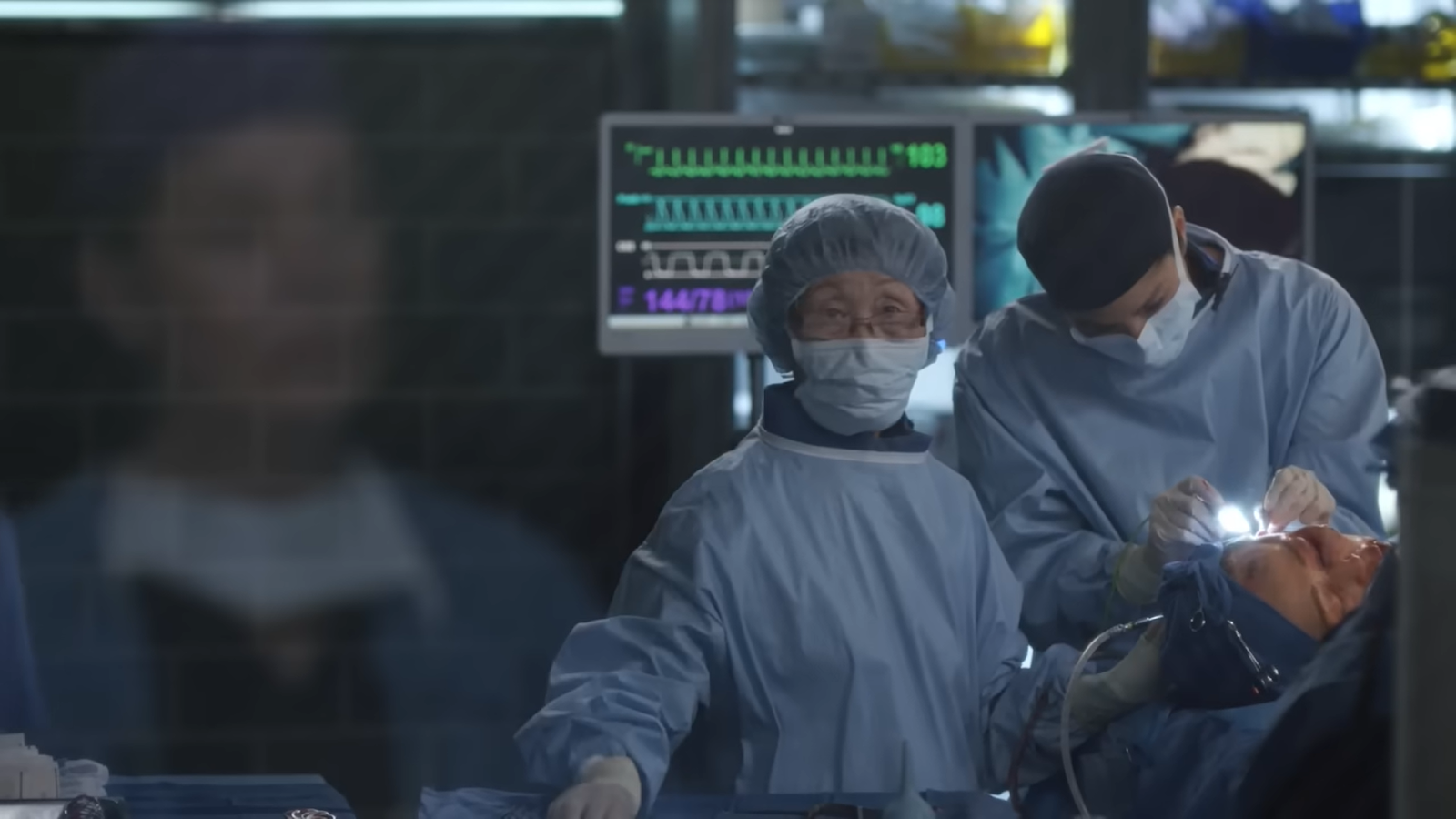 two people standing near the patient in the operating room, one stands straight and looks forward, other does surgery