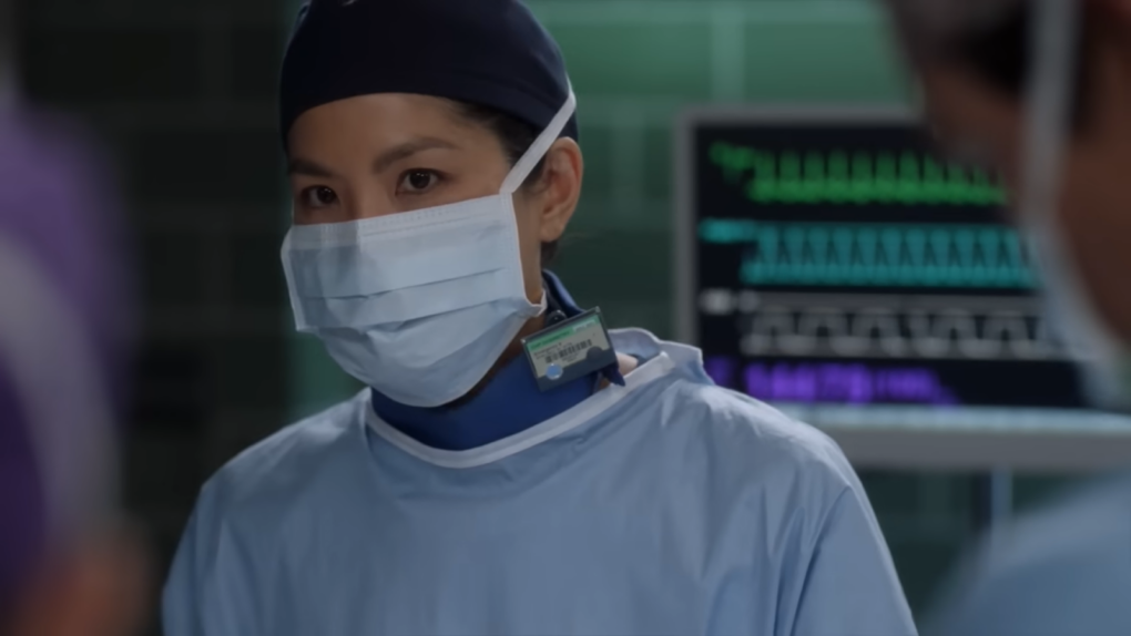 Chinese person in mask, hat, and medical suit looking forward in front of the blurred screen