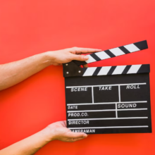 hands holding a clapperboard with words scene, take, roll on it in front of a red background