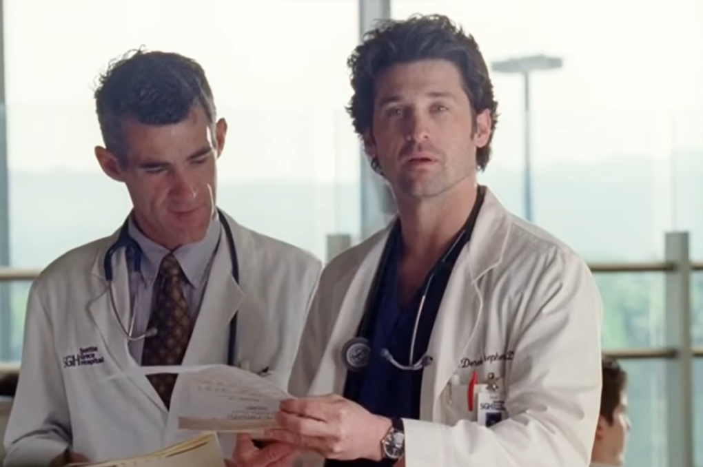 Derek Shepherd in a white medical suit and stethoscope holding papers and standing near a doctor