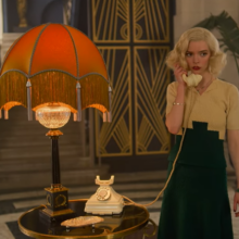 gina gray in a black and beige dress standing in the room with flowers and yellow lamp and talking on the phone