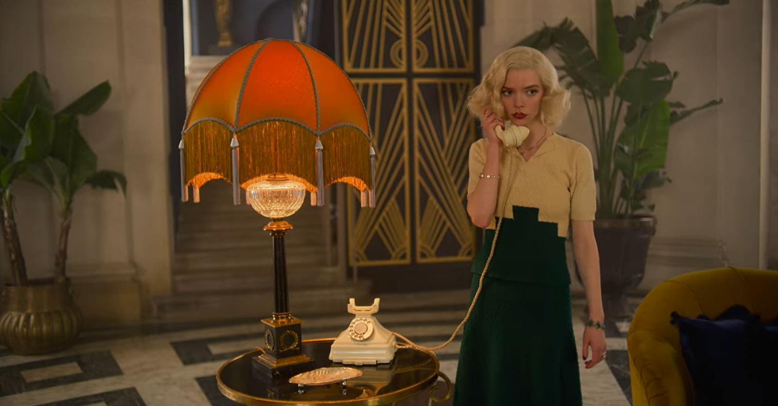 gina gray in a black and beige dress standing in the room with flowers and yellow lamp and talking on the phone