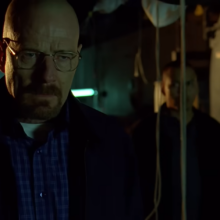 Walter White in a black jacket and blue shirt in glasses standing in semidarkness near another man who looks at him