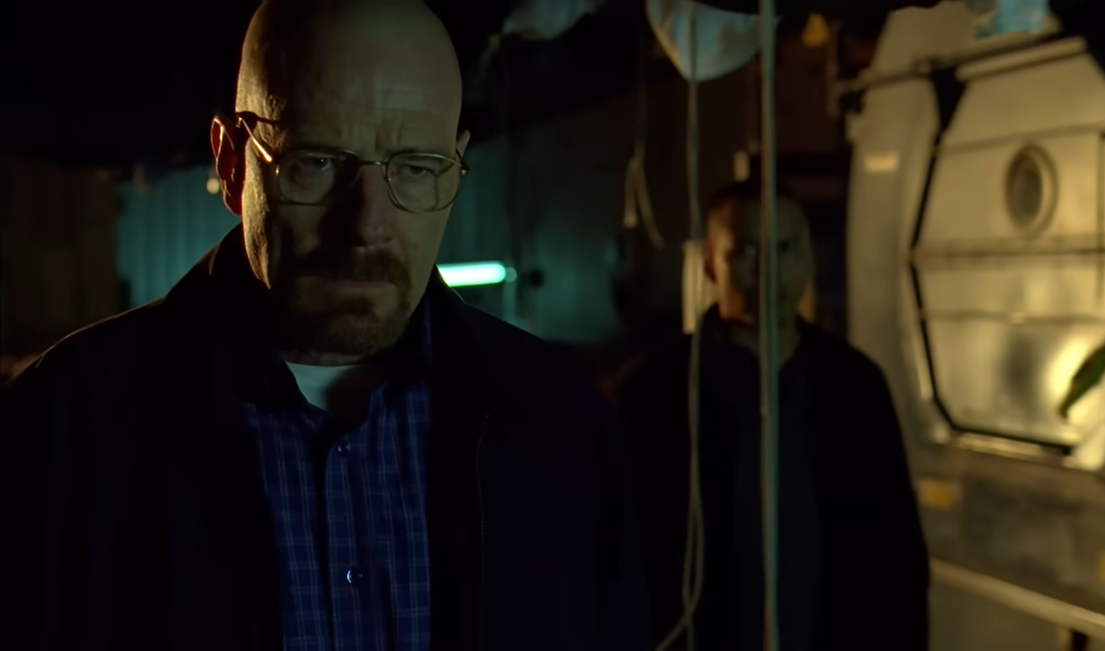 Walter White in a black jacket and blue shirt in glasses standing in semidarkness near another man who looks at him