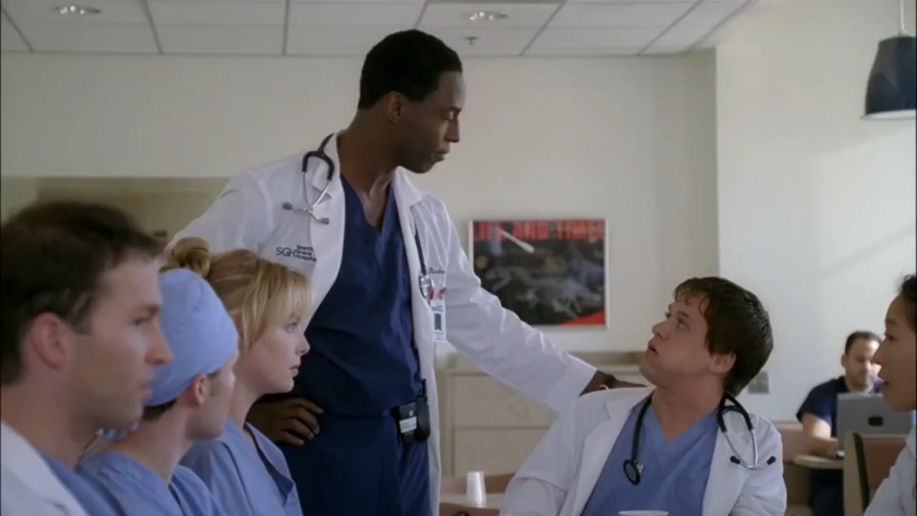 man holding hand on the shoulders and standings in front of a man with stethoscope who sits at the table, four people looking at them