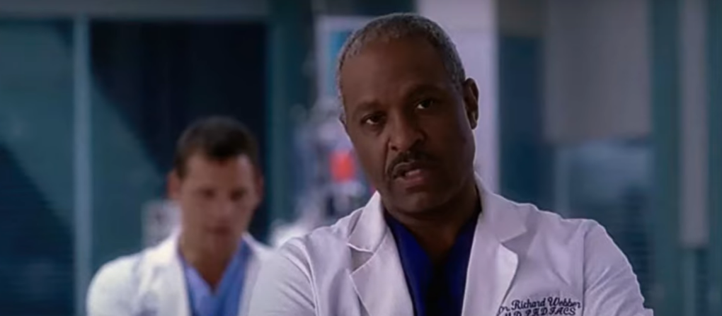 man in white medical suit and blue shirt standing straight and looking forward, other man behind him on blurred background